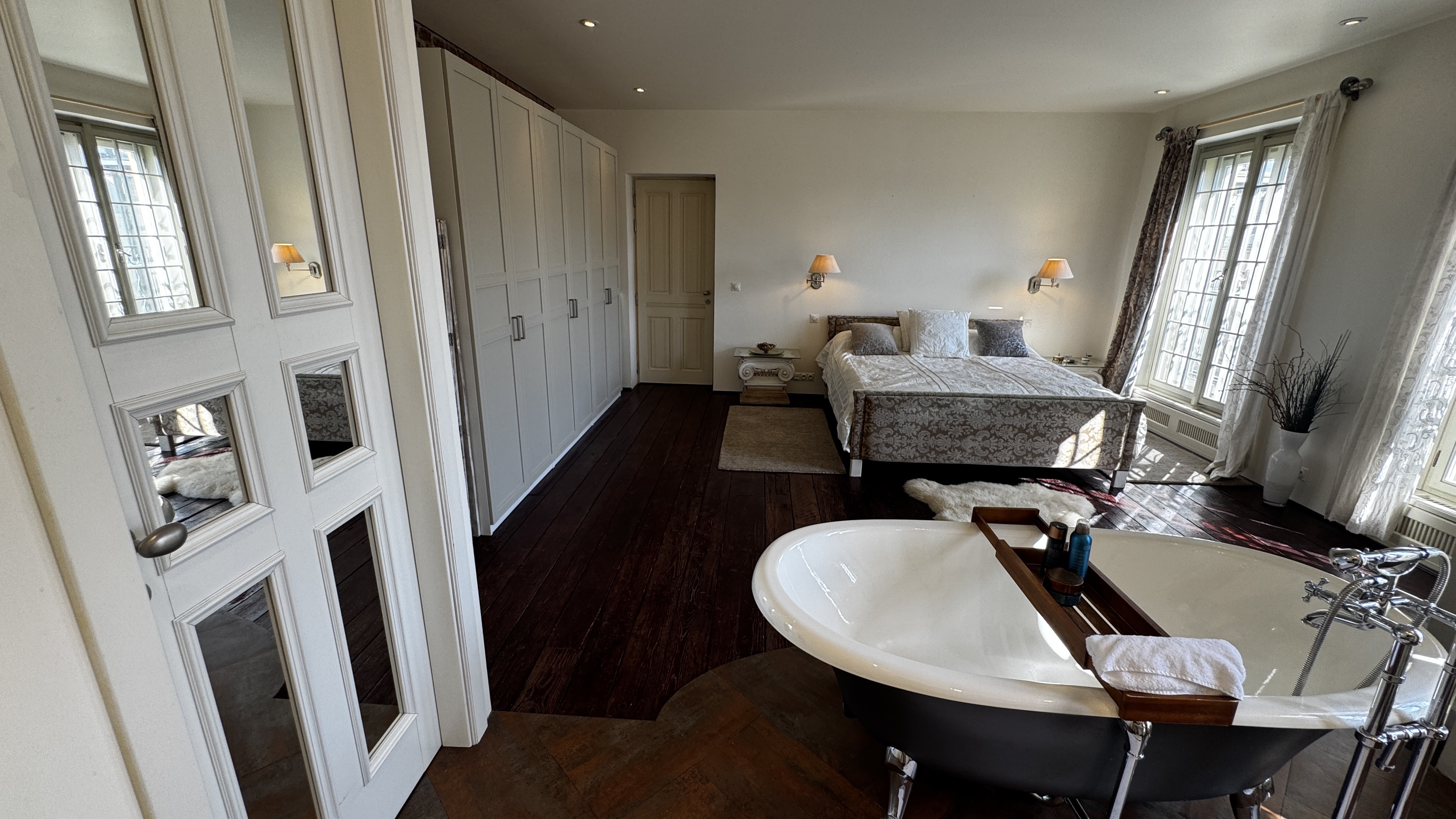 Wide view of the master sanctuary at Polska Penthouse — fitted joinery, the bed beneath tall curtained windows, and the freestanding ROCA bathtub on dark timber floors